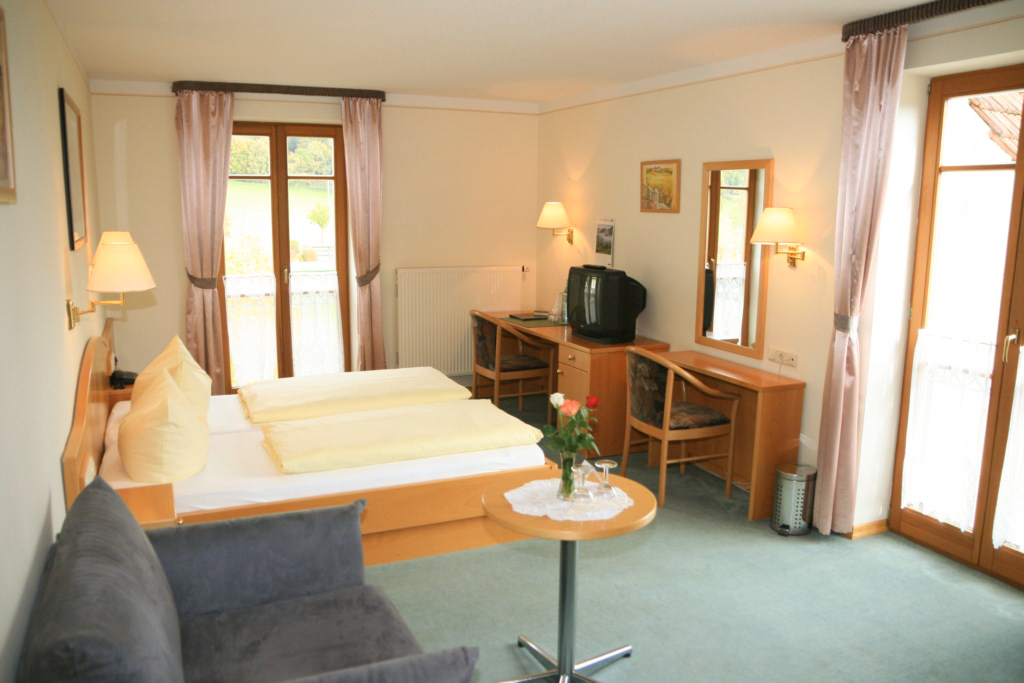 Hier ist ein Gästezimmer zu sehen. Man sieht einen hellblauen Teppich, ein Doppelbett und eine kleine Couch mit Tisch neben dem Bett. Vor dem Bett befindet sich ein Schreibtisch mit Spiegel, sowie ein Fernseher. Das Zimmer ist hell dank zwei großer Fenster.