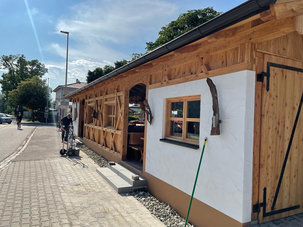 Man sieht den Biergarten von aussen. Der im Hüttenstil gebaute Biergarten hat an der rechten Seite eine weiße Mauer, eine schöne große Tür und viele Holzelemente.
