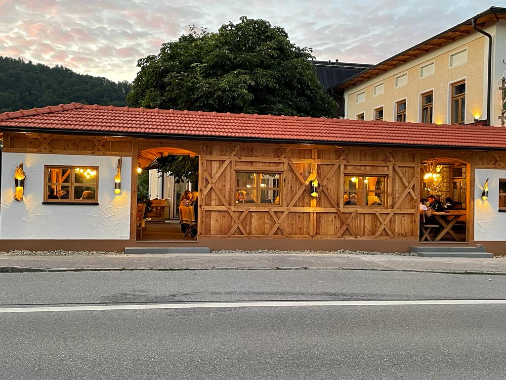 Man sieht ein zweites Bild vom Biergarten von aussen. Der im Hüttenstil gebaute Biergarten hat an der rechten Seite eine weiße Mauer, eine schöne große Tür und viele Holzelemente.